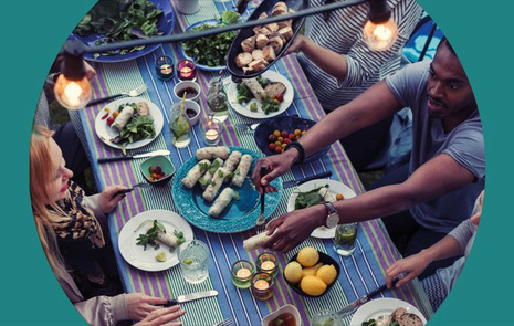 Många personer som sitter vid ett stort bord utomhus och har en vegetarisk festmåltid