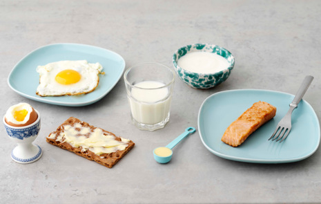 Ett ägg i en äggkopp, ett stekt ägg på ett fat, en smörgås med smör, ett glas med berikad mjölk, en matsked med matfett, en liten skål med fil, en stekt laxfilé på ett fat med en gaffel